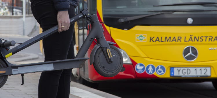 Människa står och väntar på en buss med en elsparkcykel i handen. 